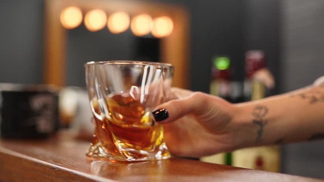 Experienced woman sommelier shaking a glass with whiskey and inhaling alcohol aroma. Female hand moves glass with expensive brandy on wooden bar counter in barbershop.