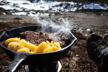 Cooking steaks and potatoes with cast iron frying pan next to sand pit at spring time.