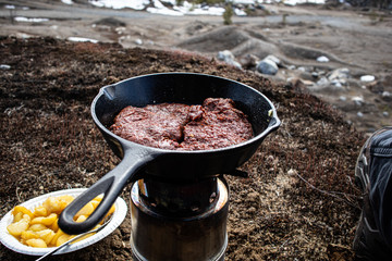 Cooking steaks and potatoes with cast iron frying pan next to sand pit at spring time.