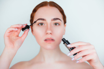 redheaded young woman looking natural applying brow mascara while doing makeup standing on isolated...