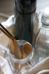 Canvas tote bag with sustainable kitchen products: stainless steel thermos bottle, glass bottle, glass jar with wooden cutlery and reusable metal straws. Selective focus, wooden background. 