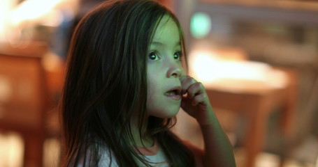 Little girl child watching TV screen at night. Small kid hypnotized staring screen scratching face.