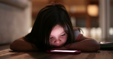 Small girl staring at cellphone screen at night. Cute kid lying on floor watching cellphone screen.