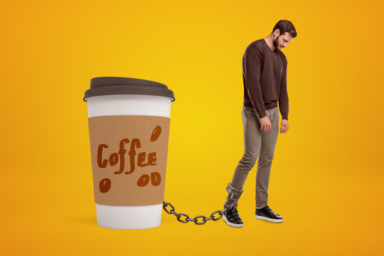 Depressed Young Man Standing And Looking Down, Chained To Huge Coffee Cup On Amber Background.