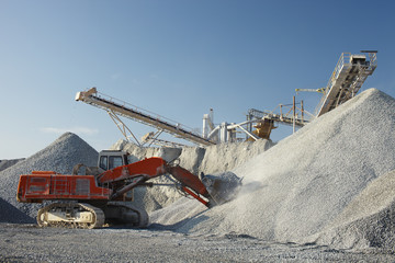 Powerful red excavator with a full bucket of crushed stone on a background of constructions of mining enterprise. Heavy mining equipment.