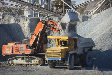 Red excavator pouring crushed stone out of a bucket into a dump truck body, closeup. Heavy mining...