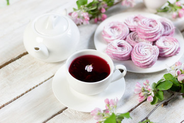 Red hibiscus tea with floating apple tree flower in white cup on wooden table with teapot, pink...