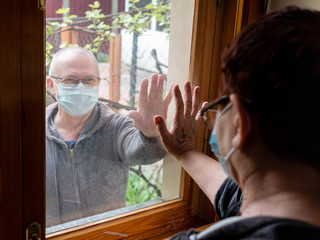 An elderly man and woman touch their palms to the window glass, unable to communicate with each other face to face due to the spread of the coronavirus.