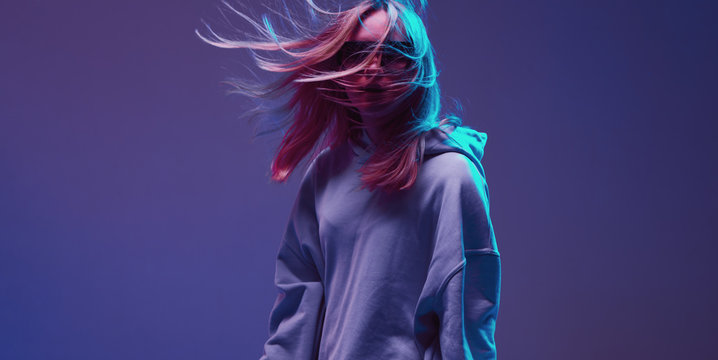 Portrait of a stylish young girl, cool posing in a hoodie, sunglasses and with developing hair, on a neon background.