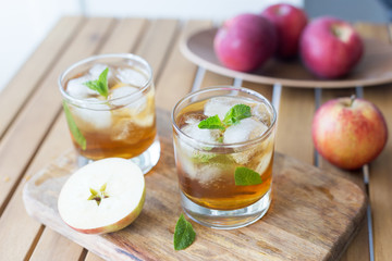 Two glass of apple juice with ice and mint at wood background. Tray with apples behind