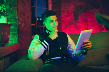 Interested. Cinematic portrait of stylish man in neon lighted interior. Toned like cinema effects, bright neoned colors. Caucasian model using gadgets, devices in colorful lights indoors. Youth