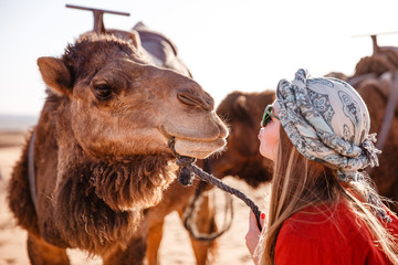 Beautiful young slim woman in a turban and sunglasses communicates with a camel at dawn in the Sahara desert. Morocco.