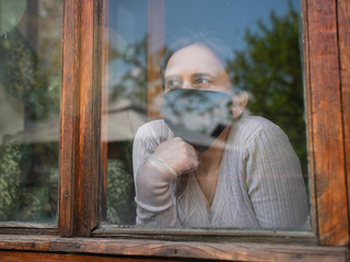 The girl in the medical mask is sad looking at the street through the window while in quarantine.