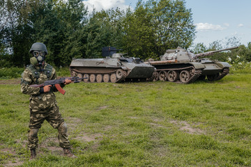 A soldier in a gas mask with a Kalashnikov rifle against the background of armored vehicles.