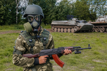 A soldier in a gas mask with a Kalashnikov rifle against the background of armored vehicles.