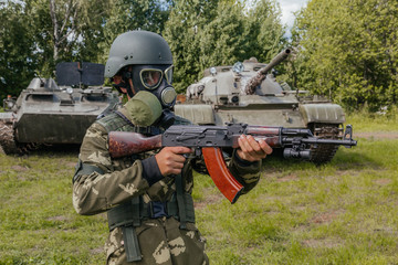 Fototapeta premium A soldier in a gas mask with a Kalashnikov rifle against the background of armored vehicles.