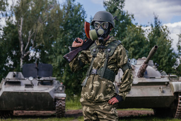 Fototapeta premium A soldier in a gas mask with a Kalashnikov rifle against the background of armored vehicles.