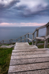 A wooden staircase leading down to a dramatic coastal seascape with ominous clouds and fog rolling in. An iceberg in the ocean off shore. North Head Trail at Signal Hill - Newfoundland, Canada.  