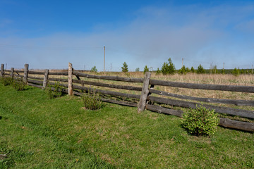 Fototapeta na wymiar Picture of a rural lanscape with old rickety wooden fence, little green bushes, grass and blue sky at spring