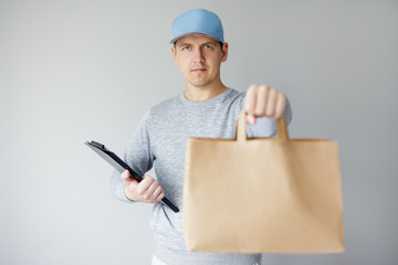 delivery concept - young delivery man holding clipboard and giving paper bag over white background