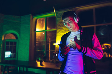 Obraz na płótnie Canvas Choosing song. Cinematic portrait of stylish man in neon lighted interior. Toned like cinema effects, bright neoned colors. Caucasian model using gadgets in colorful lights indoors. Youth culture.
