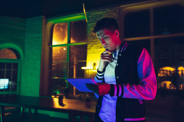 Thinking. Cinematic portrait of stylish man in neon lighted interior. Toned like cinema effects, bright neoned colors. Caucasian model using gadgets, devices in colorful lights indoors. Youth culture.