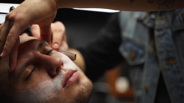 Professional barber shaves customer beard with straight razor. Beard cut with old-fashioned blade at barbershop. Handsome macho man getting his beard shaved in studio. Close-up shot.