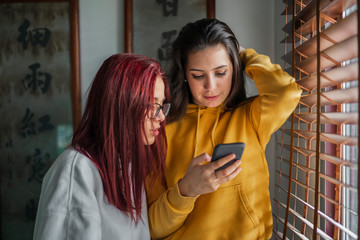 Two attractive young girls next to a window consulting the mobile phone or smartphone and laughing and talking to each other
