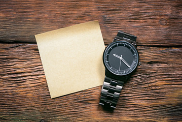 Wristwatch and blank paper sheet on brown wooden table background.