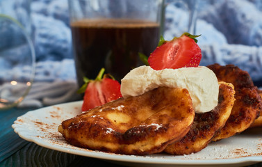Delicious and useful breakfast of cheese pancakes with sour cream and strawberries cooked at home, with strong black coffee on blue background of wood and plaid.
