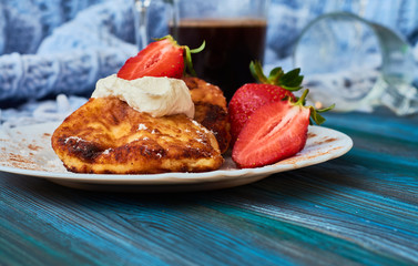 Delicious and useful breakfast of cheese pancakes with sour cream and strawberries cooked at home, with strong black coffee on blue background of wood and plaid.