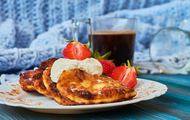 Delicious and useful breakfast of cheese pancakes with sour cream and strawberries cooked at home, with strong black coffee on blue background of wood and plaid.
