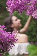 A young girl in a white blouse stands between the purple lilac flowers