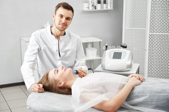 Beautician man doing anti-aging procedure of rf lifting face skin of a young woman in a white beauty parlor