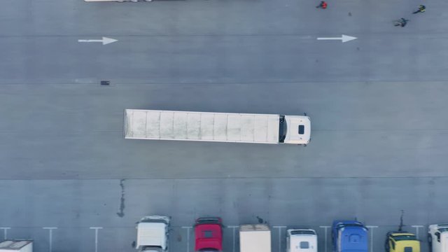 Aerial following view of the semi truck with cargo trailer is travelling on the parking lot along the warehouse of the logistics park