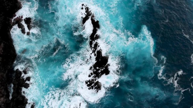 Aerial top down shot of big waves crashing into cliffs near the ocean shore of Porto Moniz, Madeira island, Portugal. Drone flying over blue turquoise water