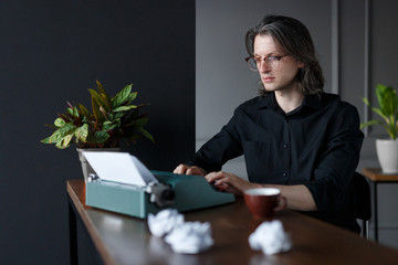 Portrait of a young man, seated at the table, working on typewriter. Shooting in studio, over gray background. Copy space. Horizontal view. - Powered by Adobe