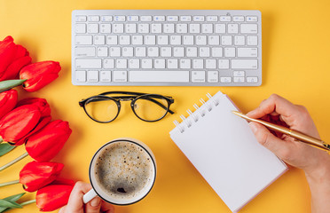 Home office workplace: cup of coffee, empty notebook, keyboard, tulip flowers on yellow background and female hand. Flat lay, top view. Work from home
