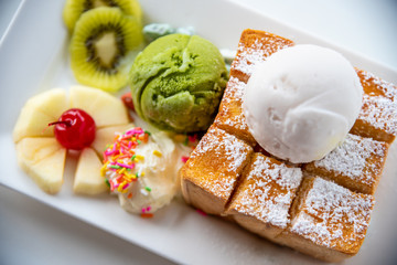 Closeup top view Honey Toast bread served with green tea and coconut milk ice cream scoop, decorate with cherry, apple, kiwi fruit, and cream sugar sprinkle on a white dish sweet cool snack in summer