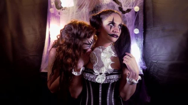 Girl with Halloween makeup is sitting in front of a mirror. Makeup of the actor. The girl in the image of the devil's Bride comes up and bites the neck of the clown girl.