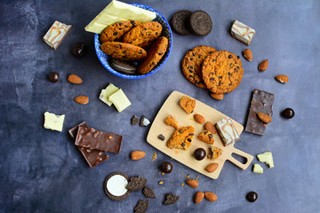 Top view on a blue bowl with dark, white chocolate and cookies and a wooden board with almonds and cookie crumbs on a dark gray background. Sweets, pastries and desserts