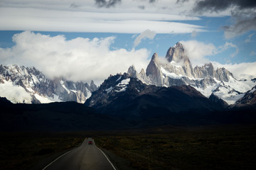 Road with a red car and the peaks of a rocky and snowy mountain on the horizon. Mountains of Fitz Roy in Argentina