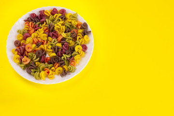 colorful raw pasta with vegetable ingredients, carrots, beets, spinach, tomatoes in a white plate on a yellow background.