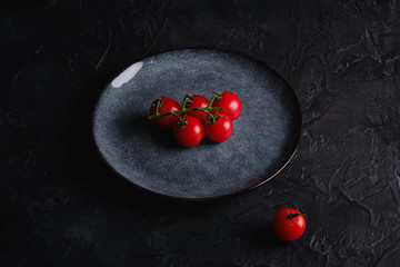 Cherry tomato branch, fresh ripe organic vegetables in grey plate on dark black textured background, angle view