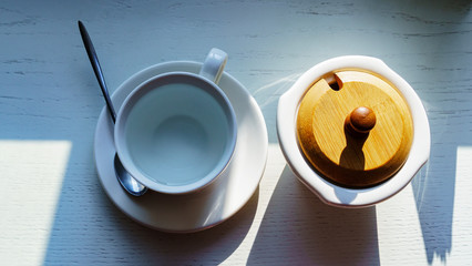A cup of hot water for tea and a sugar bowl stand on a white wooden table in the rays of lighting.