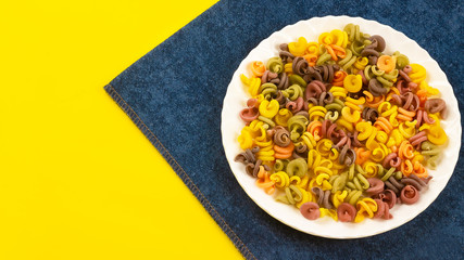 colorful raw pasta with vegetable ingredients, carrots, beets, spinach, tomatoes in a white plate on a yellow background and a blue napkin.