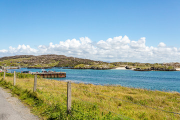 Fototapeta na wymiar Seafront promenade with wire fence with bay, pier and rocky hills in background, sunny spring day on Inishbofin Island, County Galway, Ireland