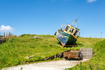 Old and abandoned boat on a hill with green grass next to an open car trailer on a farm, sunny...