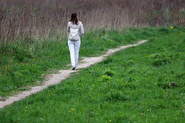 Slim woman in white jeans walking on a path on green meadow. Concept of spring season, vacation in countryside, enjoying the nature
