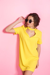 Beautiful girl wearing yellow dress and sunglasses posing on pink background in studio. Looking away of camera.